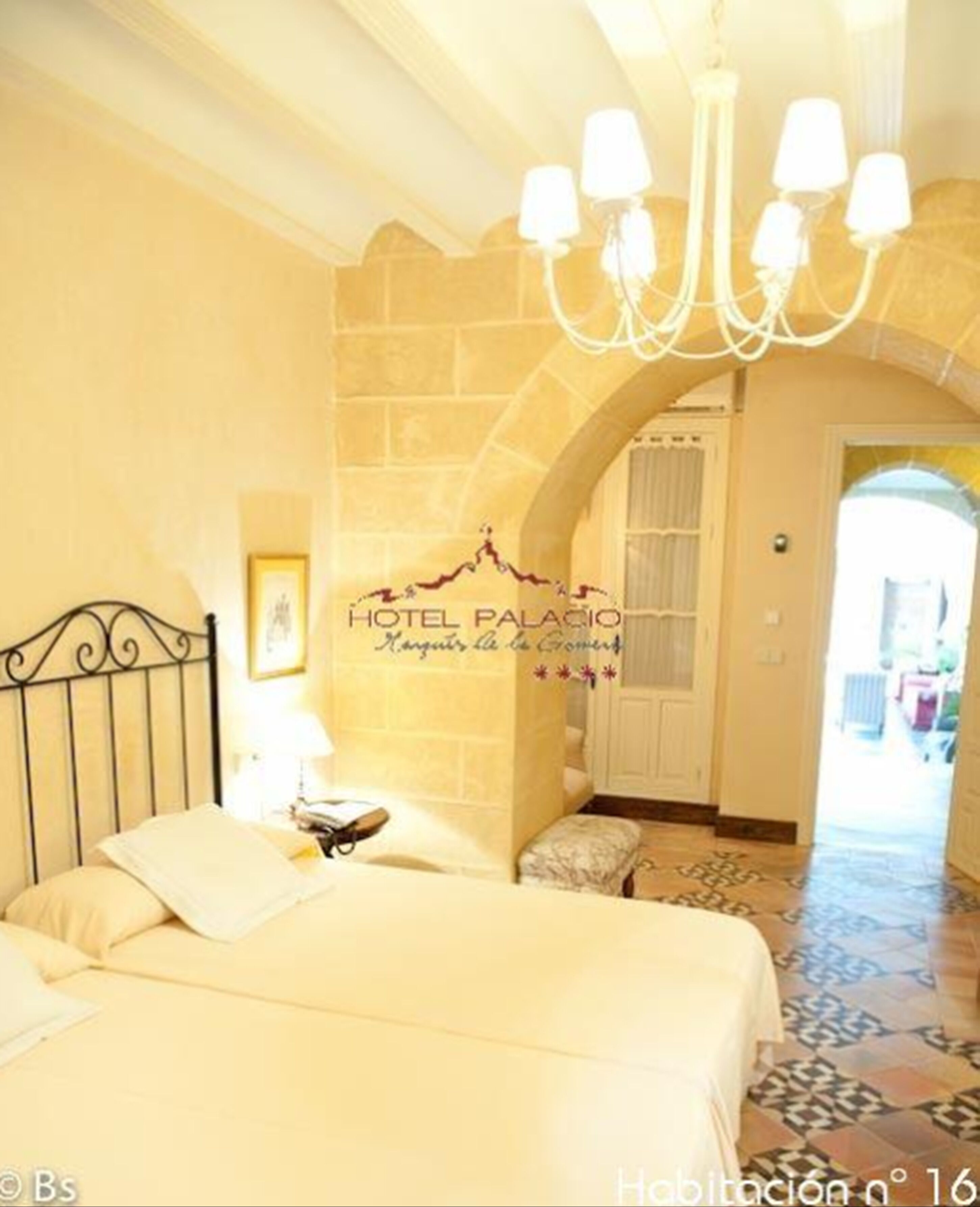 Una habitación de hotel con cama, candelabro, pared de piedra y puerta que da a otra estancia.