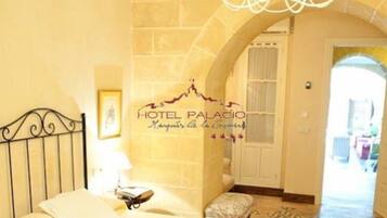 Una habitación de hotel con cama, candelabro, pared de piedra y puerta que da a otra estancia.