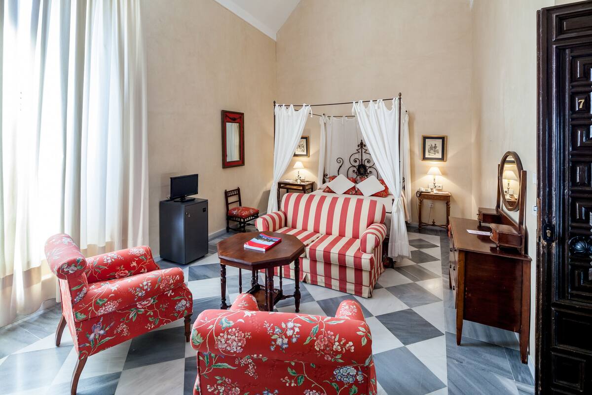 Ein Zimmer mit Schachbrettmuster-Boden, einem Blumen-Sessel, einer gestreiften Couch, einem Bett mit Baldachin, einem Schreibtisch und einem Kleiderschrank.