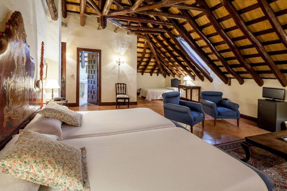 Ein Schlafzimmer mit einem großen Bett, zwei blauen Sesseln, einer Holzbalkendecke und einem kleinen Tisch mit Lampe.
