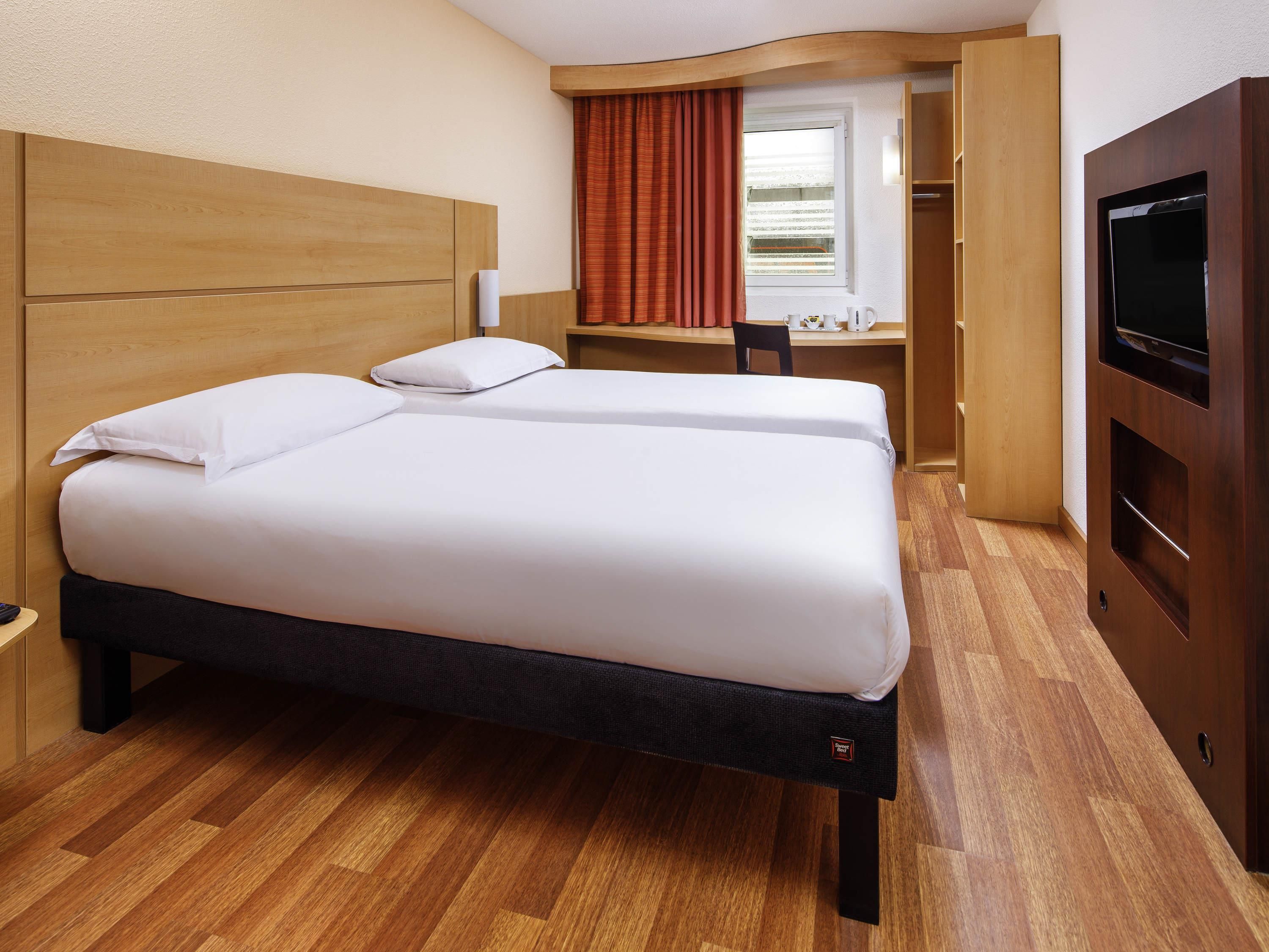 A hotel room with a wooden headboard, a bed with white bedding, a desk with a chair, and a television.