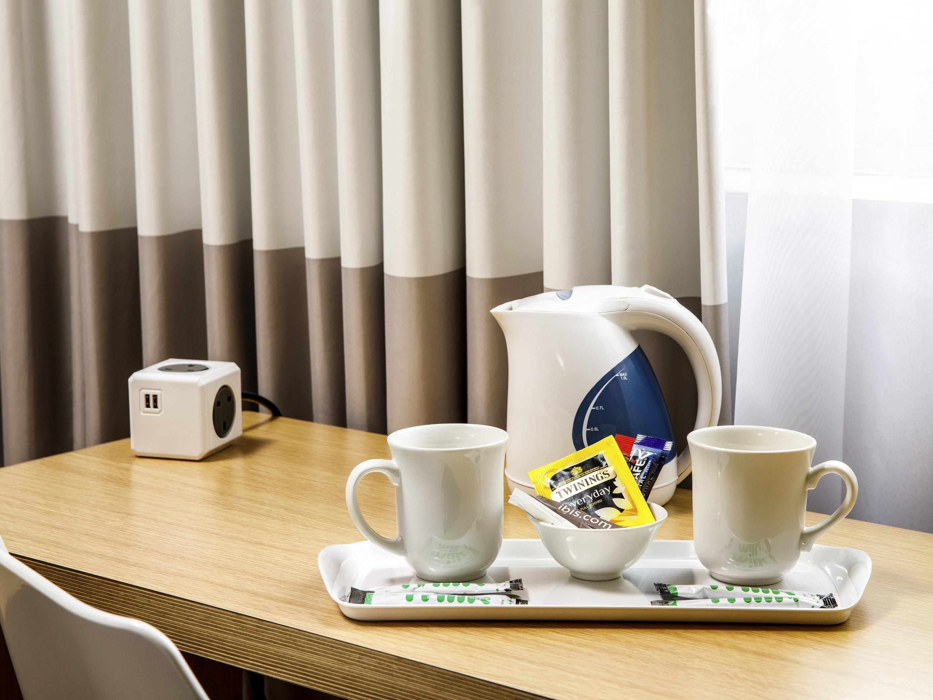 Hotellihuone, jossa on puinen pöytä, kaksi valkoista kuppia, tarjotinalusta, jossa on teekannu ja Twinings-teepaketteja, sekä pistorasiarakenne.