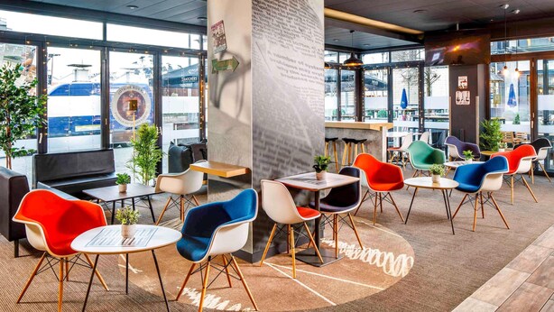 A modern cafe with colorful chairs, a central pillar with text, and large windows offering a view of the street. - ibis Bristol Centre (Bristol)