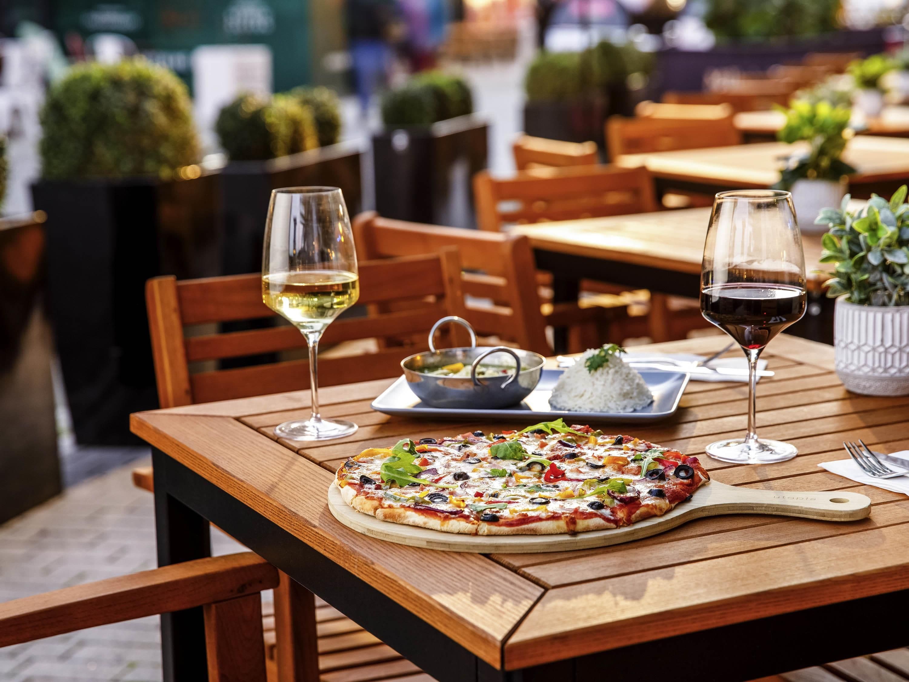 A wooden table with a pizza, two glasses of wine, a bowl of soup, and a plate of rice.