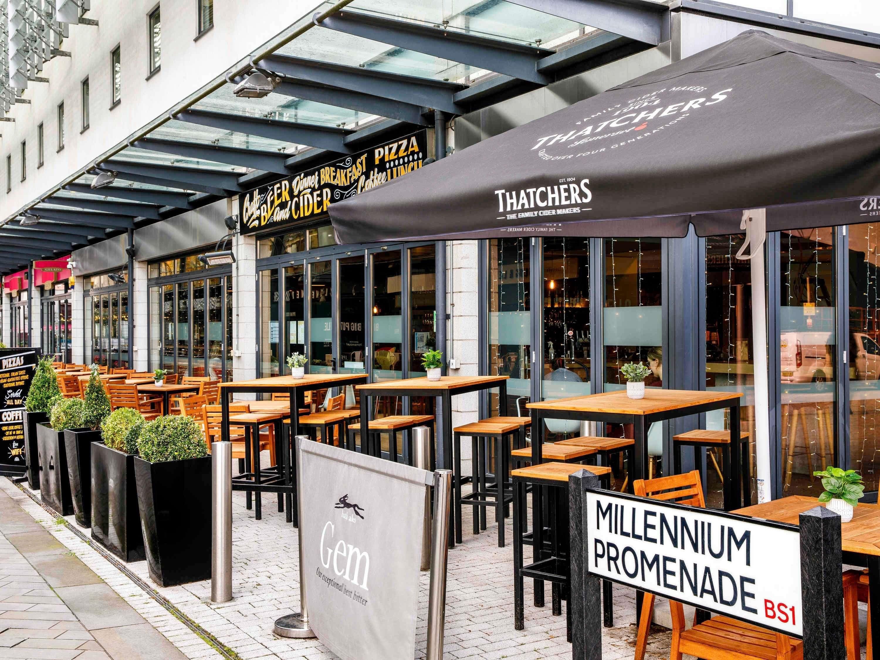 Sitzbereich im Freien mit Tischen und Stühlen, ein Schild für 'Gem' und ein Schild für die 'Millenium Promenade'.