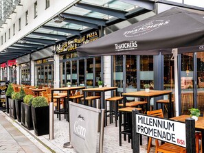 Outdoor seating with tables and chairs, a sign for 'Gem,' and a 'Millenium Promenade' sign. - ibis Bristol Centre (Bristol)