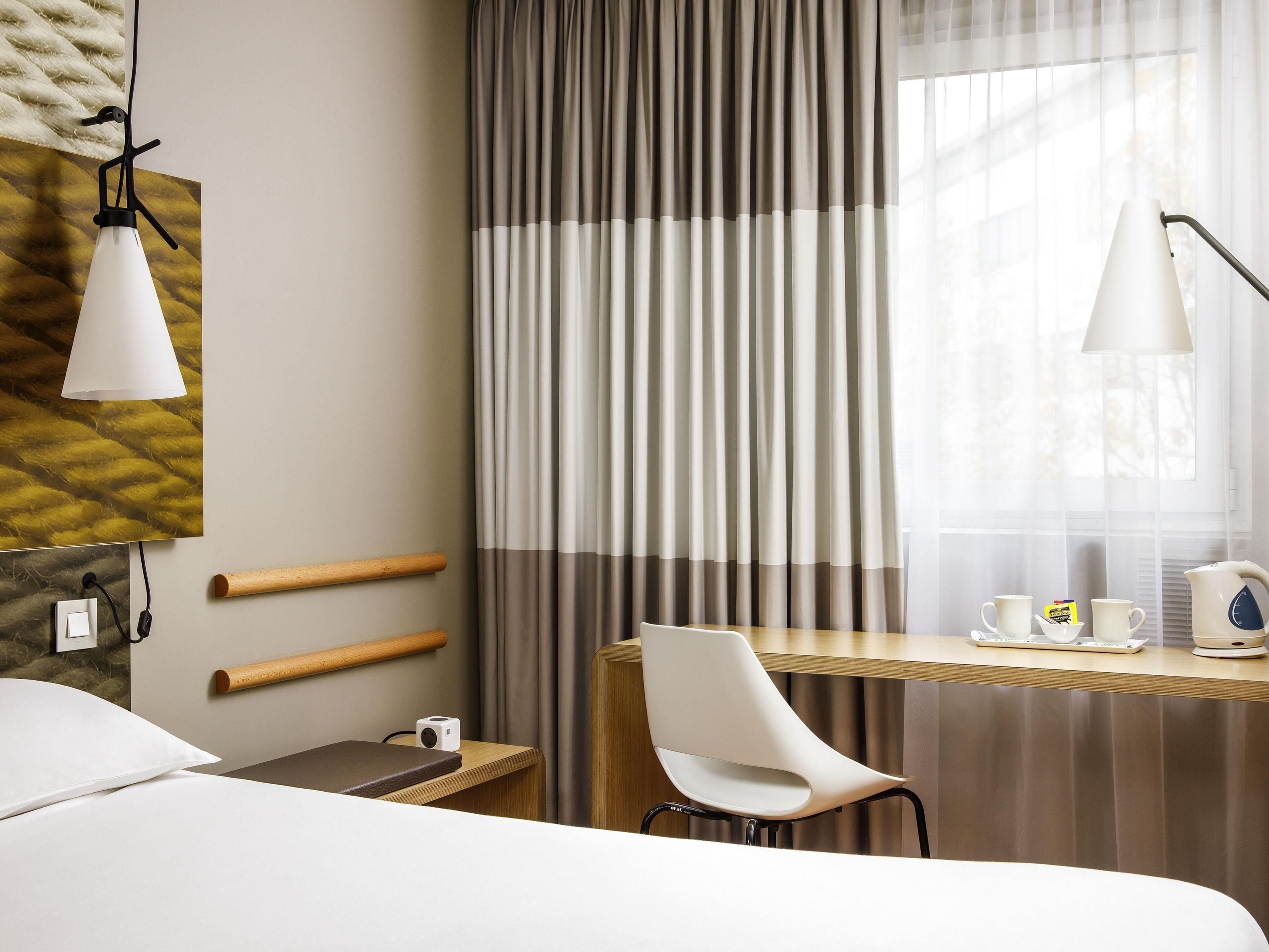 A hotel room with a bed, a desk with a chair, a lamp, a coffee maker, and curtains.