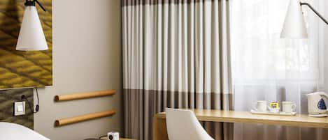 A hotel room with a bed, a desk with a chair, a lamp, a coffee maker, and curtains.
