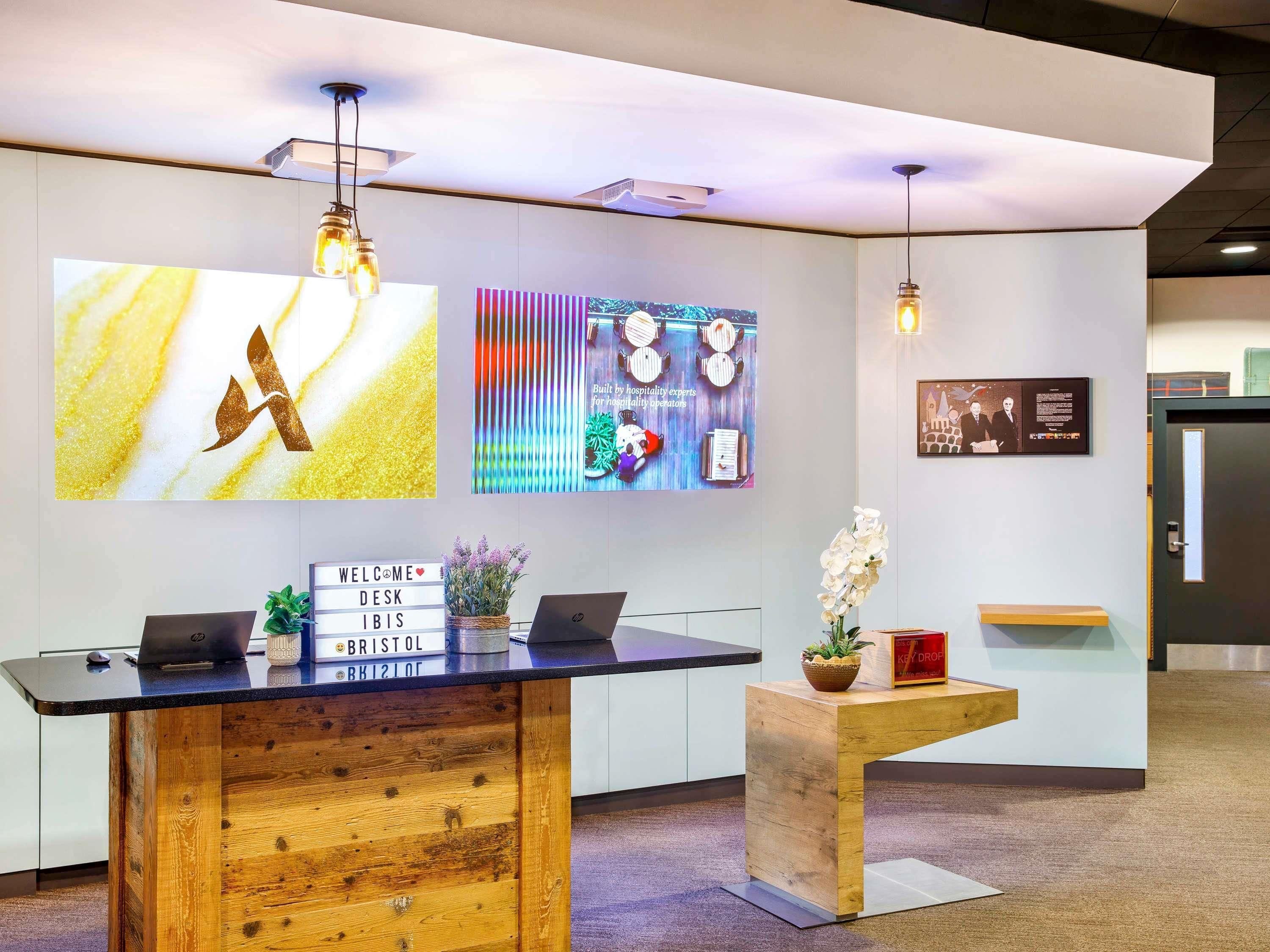 A reception desk with a sign reading 'WELCOME DESK IBIS,' a laptop, and a potted plant.
