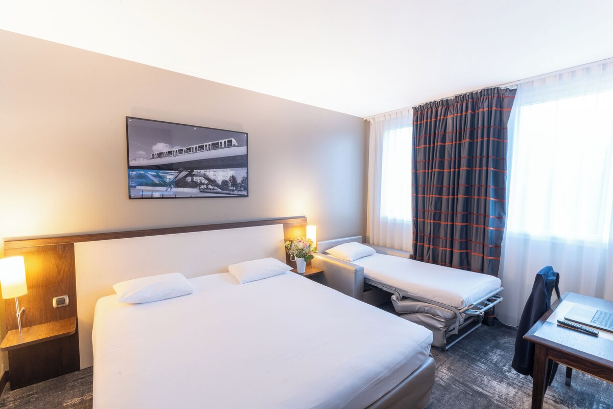 A hotel room with two beds, a desk, a chair, a lamp, a picture on the wall, and a window with curtains.