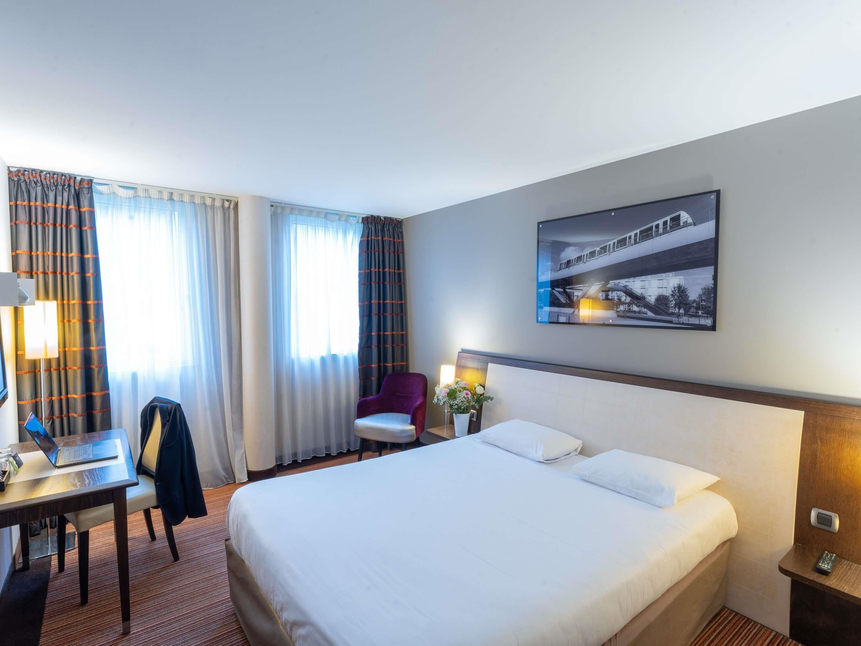 Habitación de hotel con cama, escritorio con una laptop, una silla, mesita de noche con un teléfono y un cuadro colgado en la pared.
