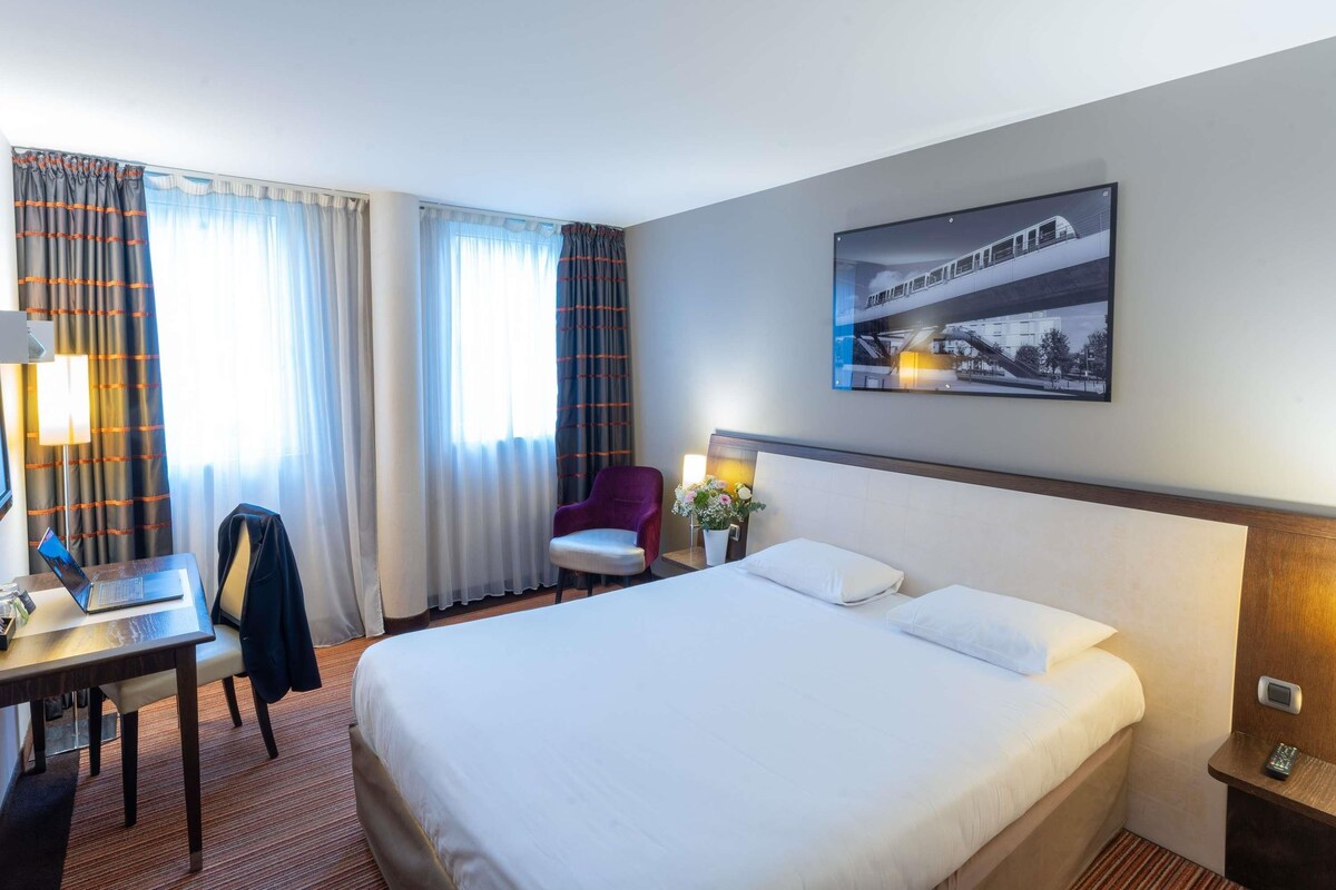 Habitación de hotel con cama, escritorio con una laptop, una silla, mesita de noche con un teléfono y un cuadro colgado en la pared.