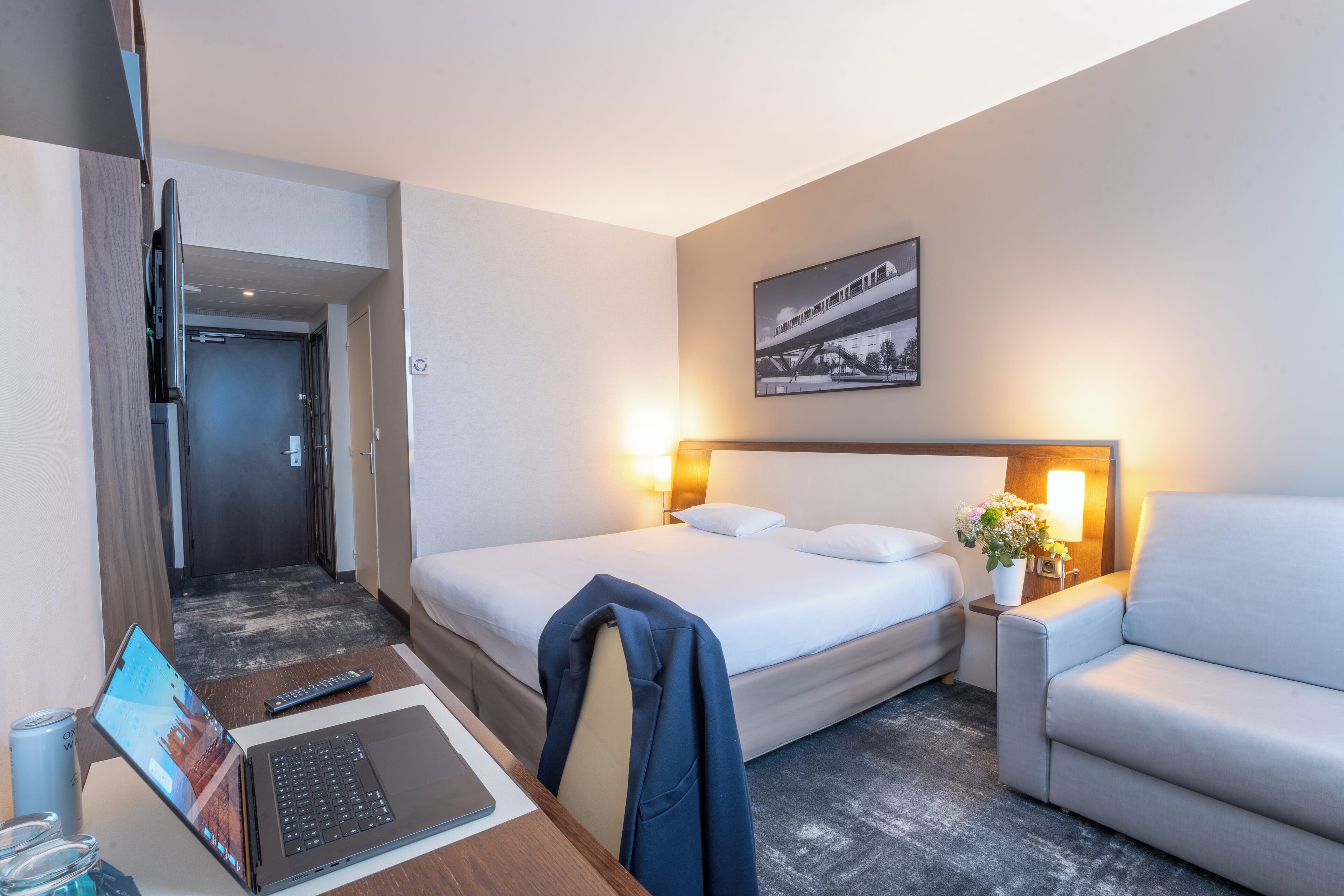 A hotel room with a bed, a desk with a laptop, a chair, a bedside table with a lamp, and a framed picture on the wall.