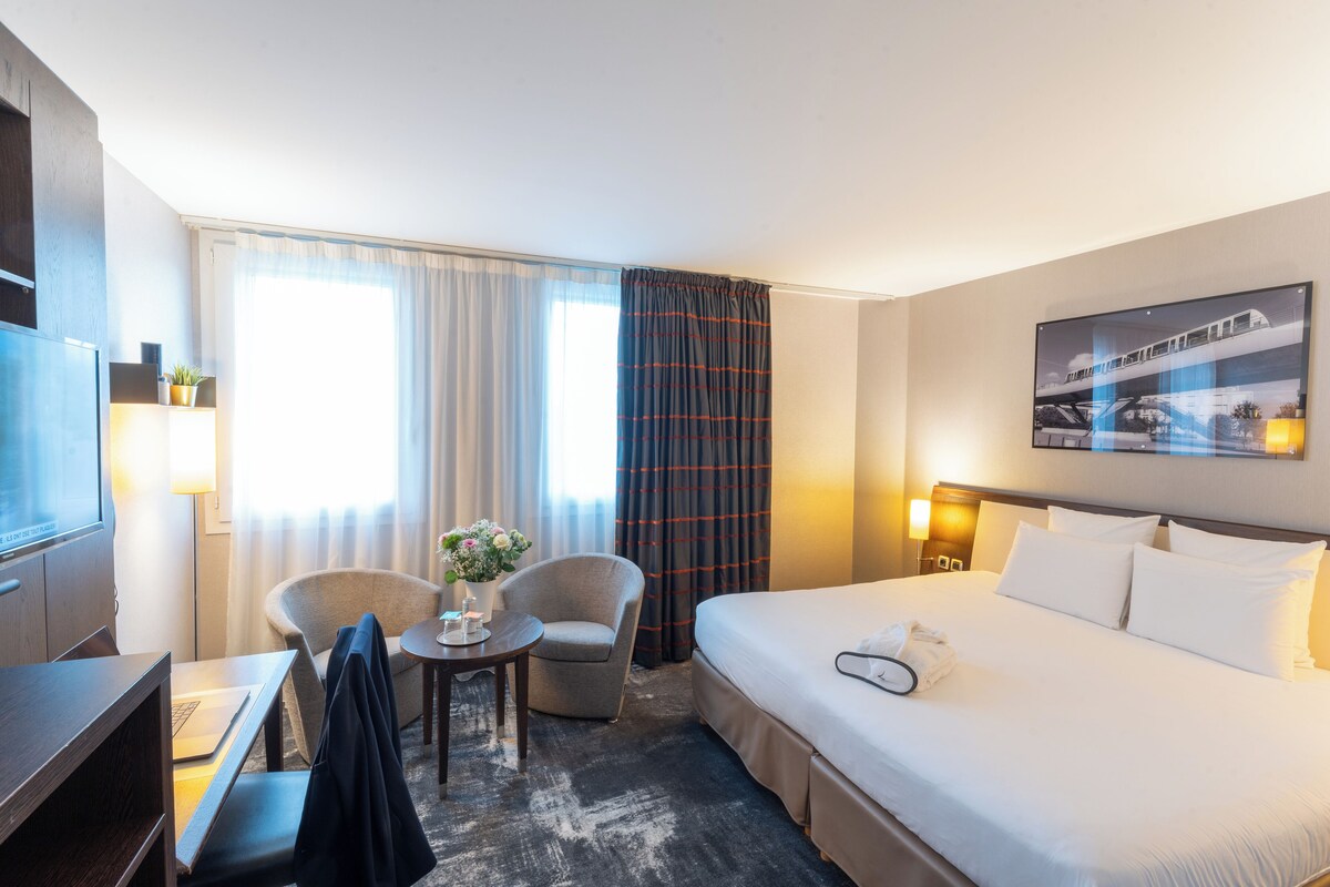 A hotel room with a bed, a desk, a chair, a small table with a vase of flowers, and a window with curtains.