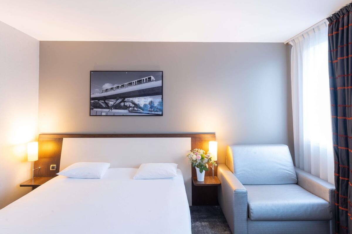 Quarto de hotel com cama, duas mesas de cabeceira com abajures, uma cadeira e um quadro pendurado na parede.