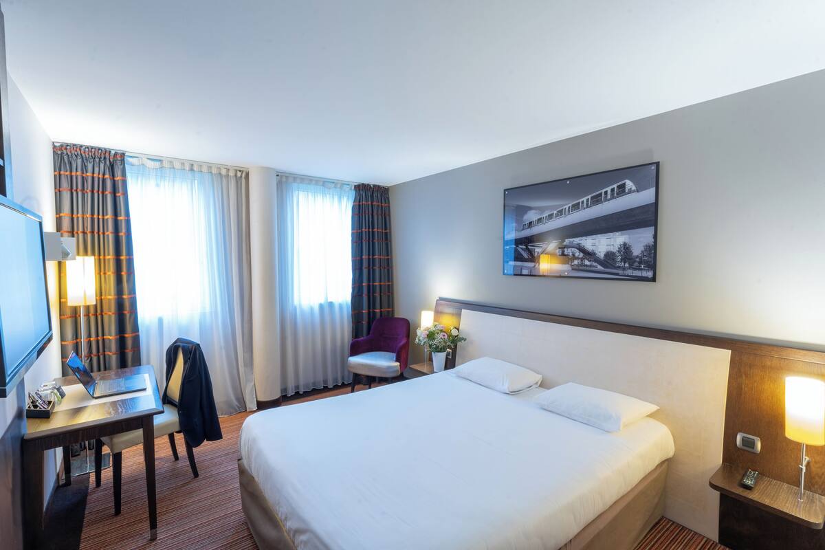 Quarto de hotel com cama, escrivaninha, cadeira e televisão na parede.