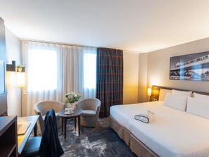 A hotel room with a bed, two chairs, a small table with a vase, a desk with a laptop, and a wall-mounted TV.