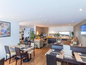 A modern hotel dining area with a central kitchen, multiple seating options, and a television screen.