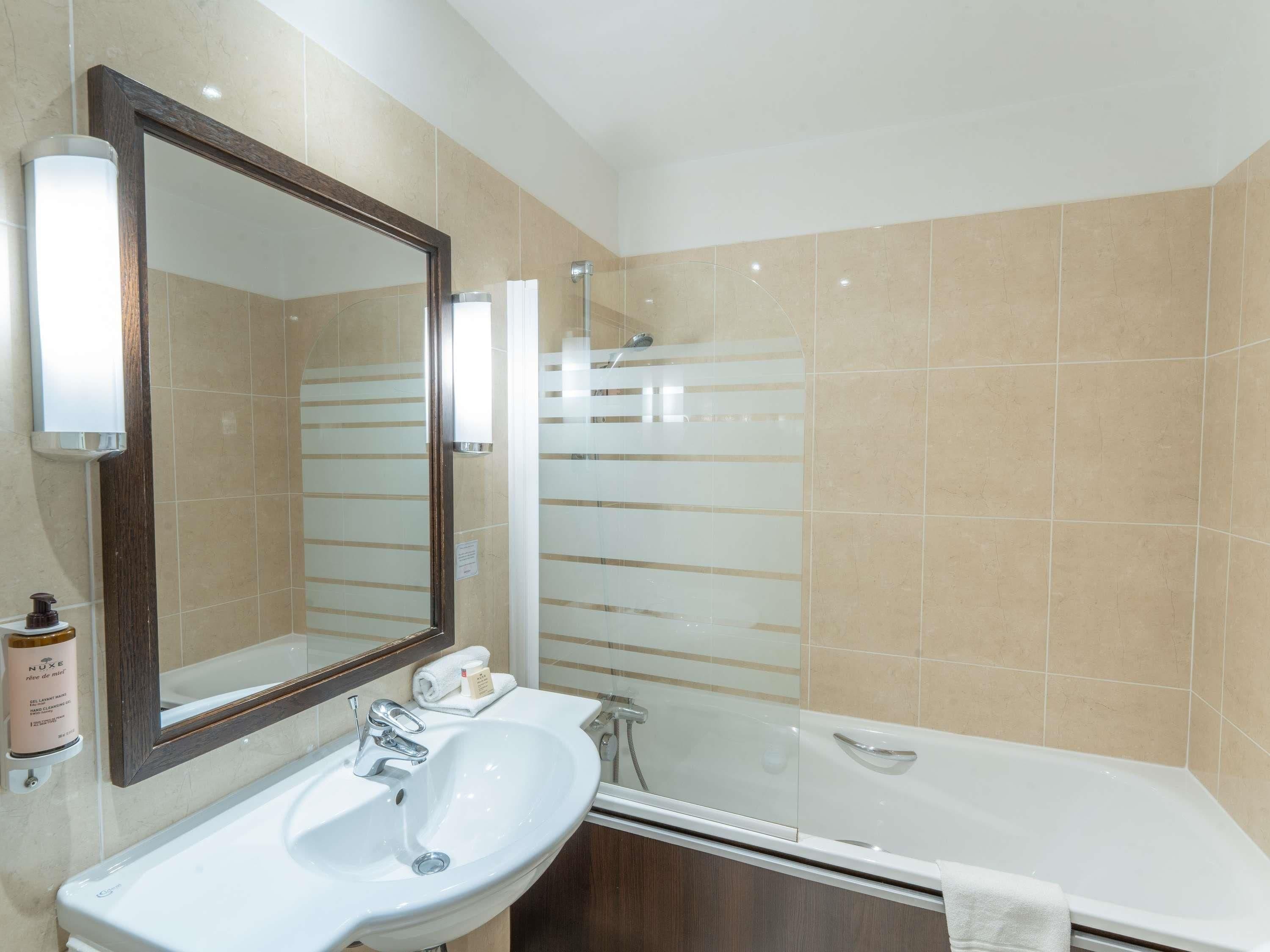 Une salle de bain avec un lavabo blanc, un grand miroir, une cabine de douche en verre et des murs recouverts de carreaux beiges.