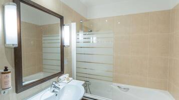 Une salle de bain avec un lavabo blanc, un grand miroir, une cabine de douche en verre et des murs recouverts de carreaux beiges.