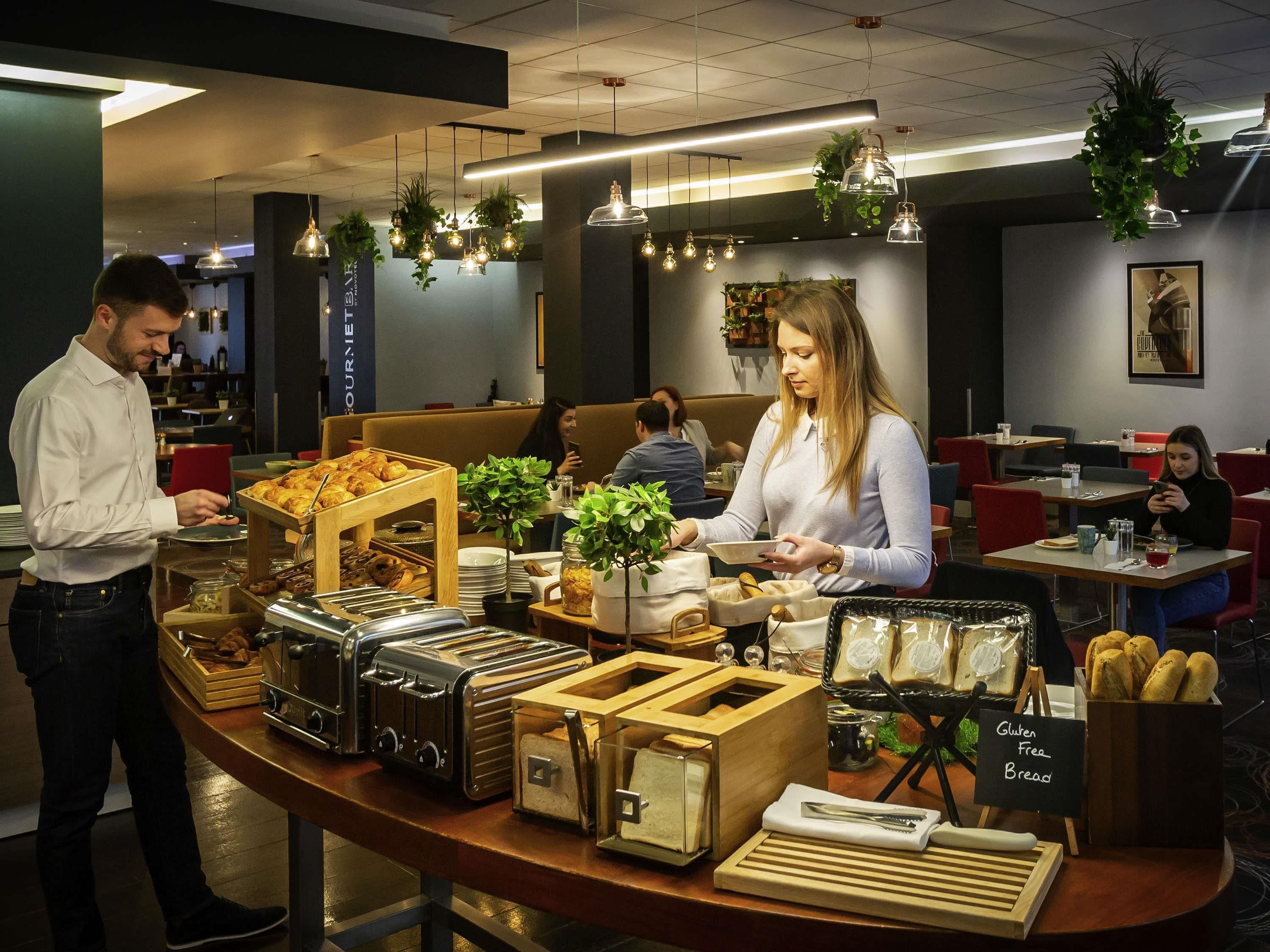 A buffet area with various food items, a sign indicating gluten-free options, and a woman serving food.