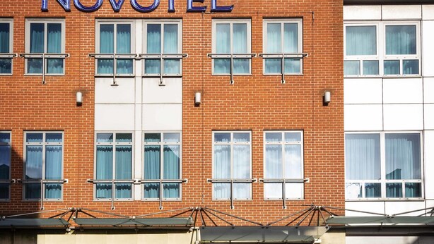 A brick building with the Novotel logo, multiple windows, and a sign indicating it is a hotel.