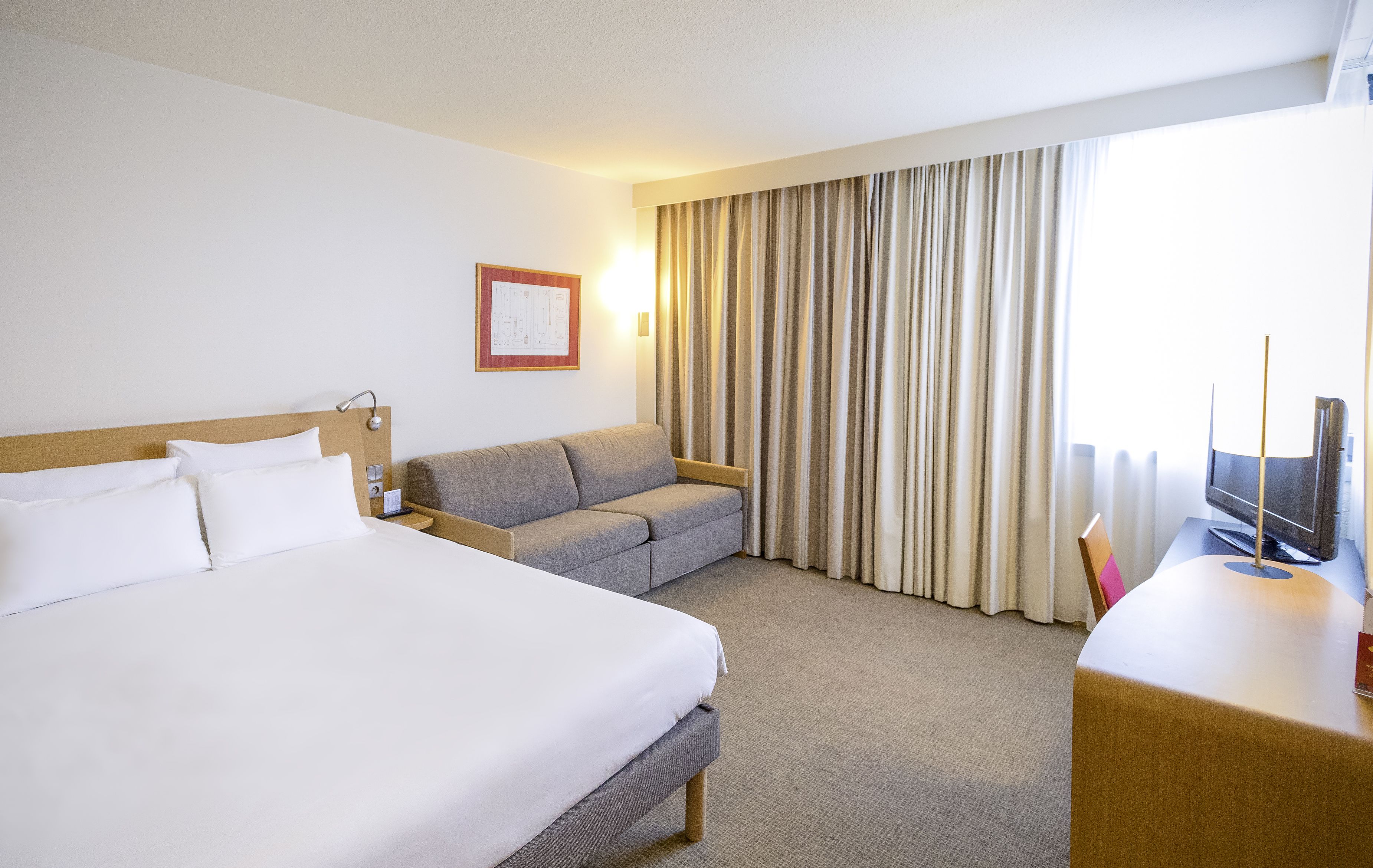 Habitación de hotel con una cama grande, un sofá, un escritorio con televisor y una ventana con cortinas.