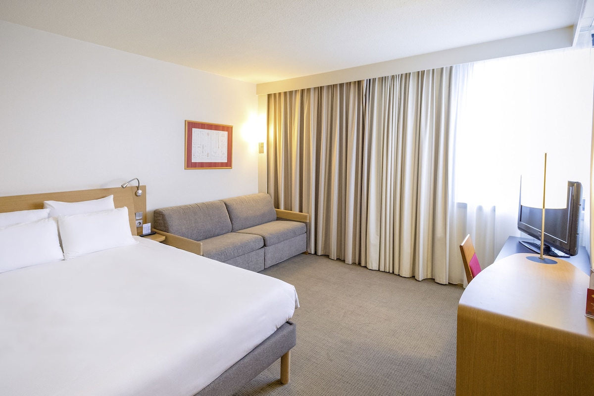 A hotel room with a large bed, a sofa, a desk with a television, and a window with curtains.