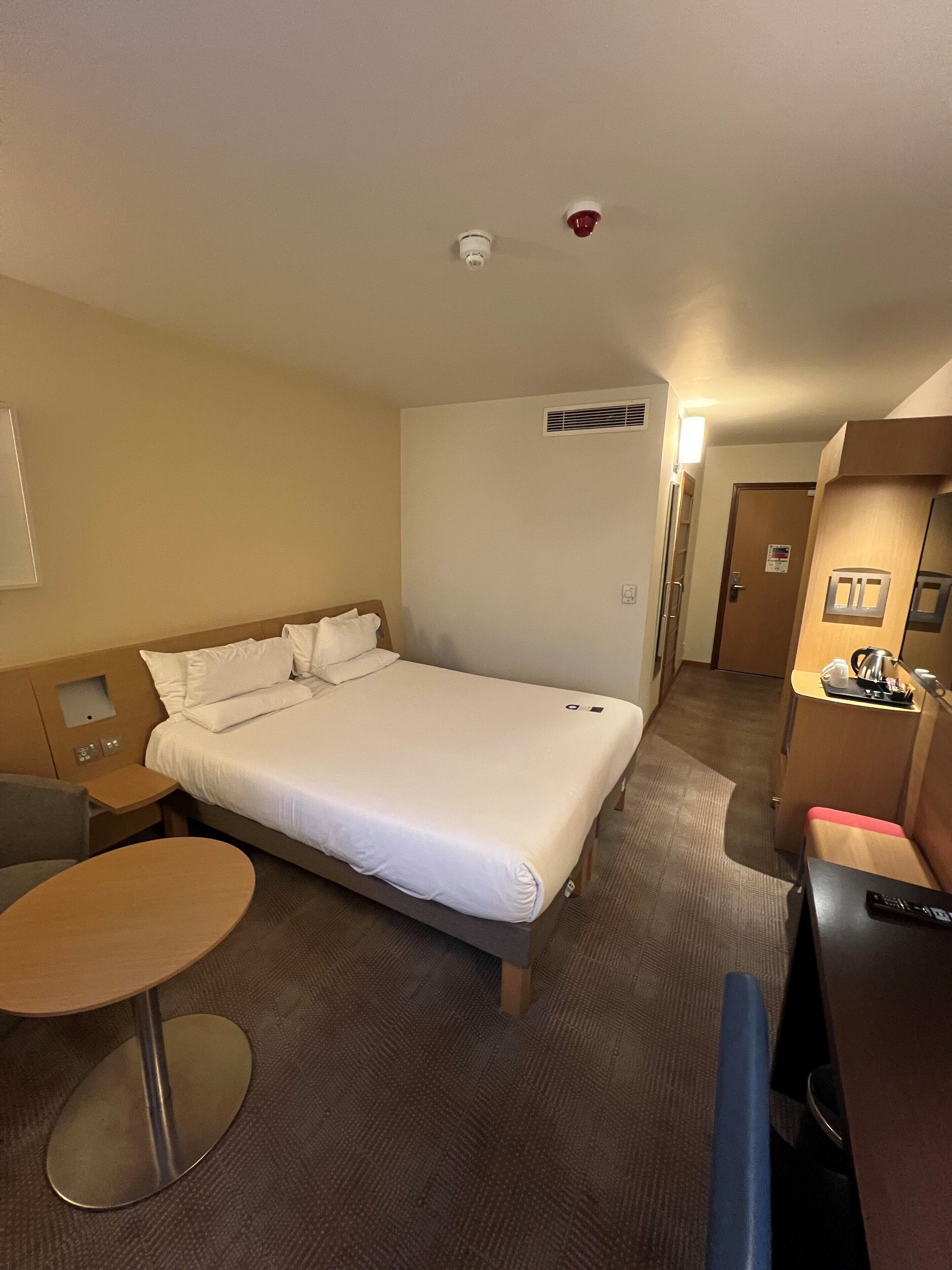 A hotel room with a large bed, a small table, a desk, and a kitchenette.