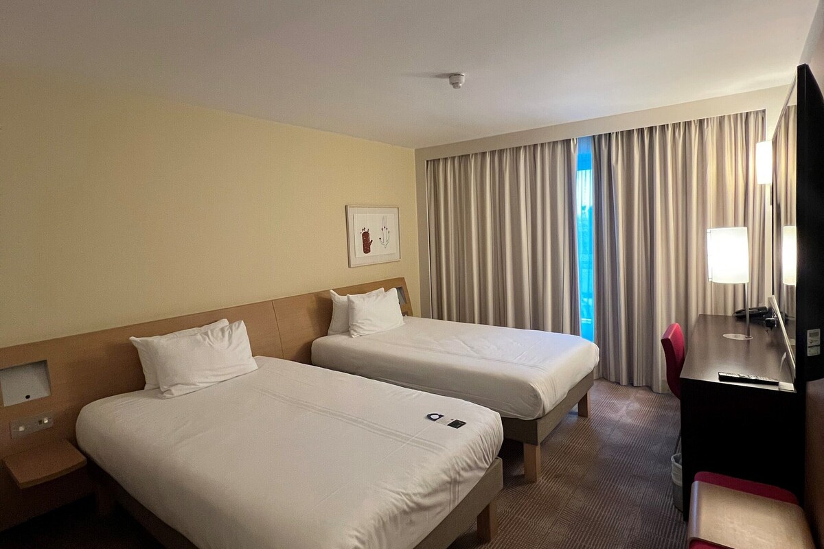 A hotel room with two beds, a desk, a chair, a TV, and a window with curtains.