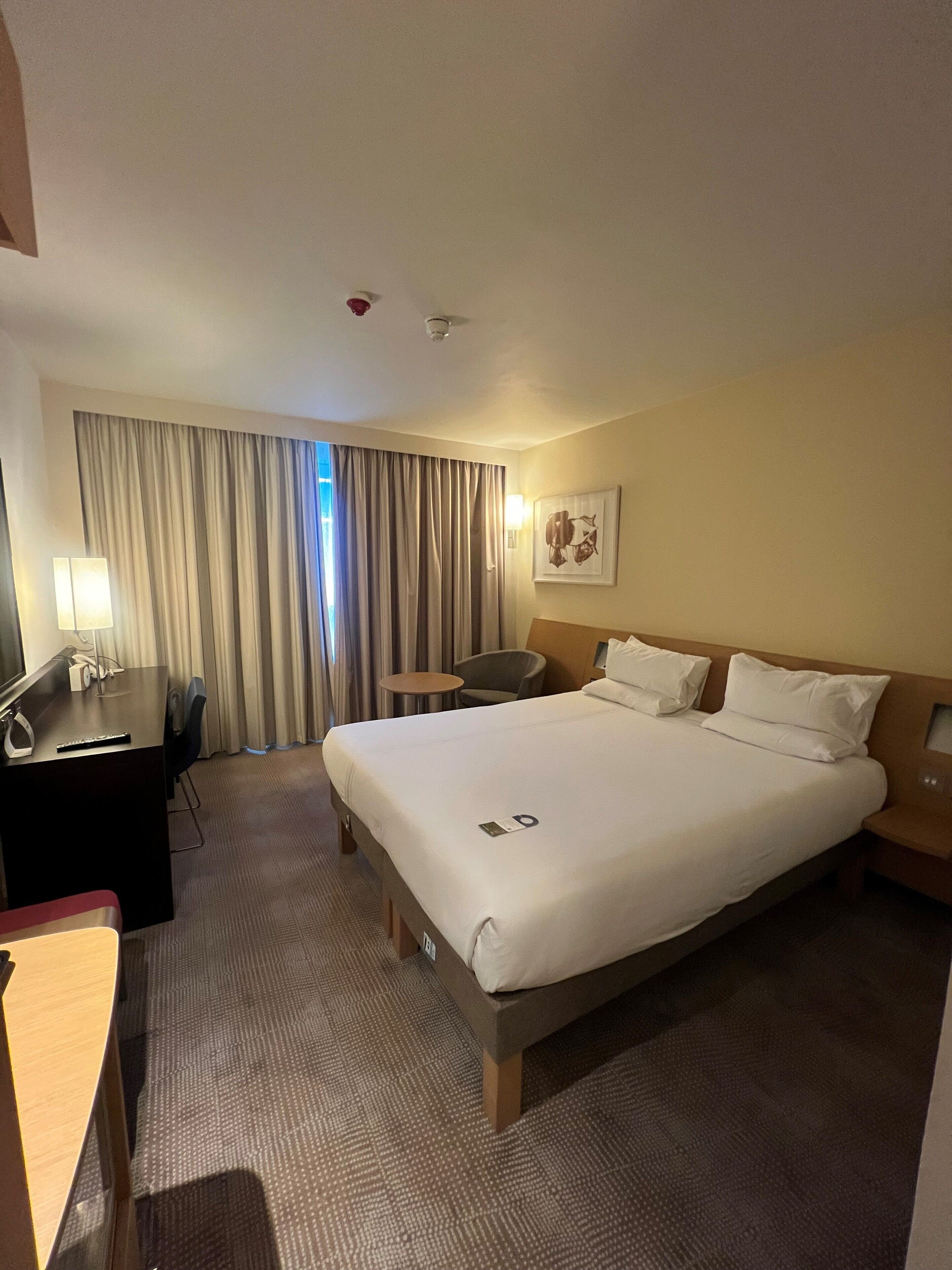 A hotel room with a large bed, a desk with a computer, a chair, and a small table.