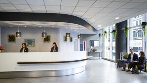 Una moderna área de recepción con un mostrador curvo, dos recepcionistas y un espacio de espera con dos huéspedes.