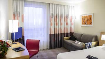 A hotel room with a bed, a desk with a chair, a sofa, a TV, and a window with curtains.
