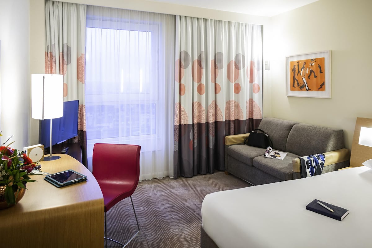 A hotel room with a bed, a desk with a chair, a sofa, a TV, and a window with curtains.