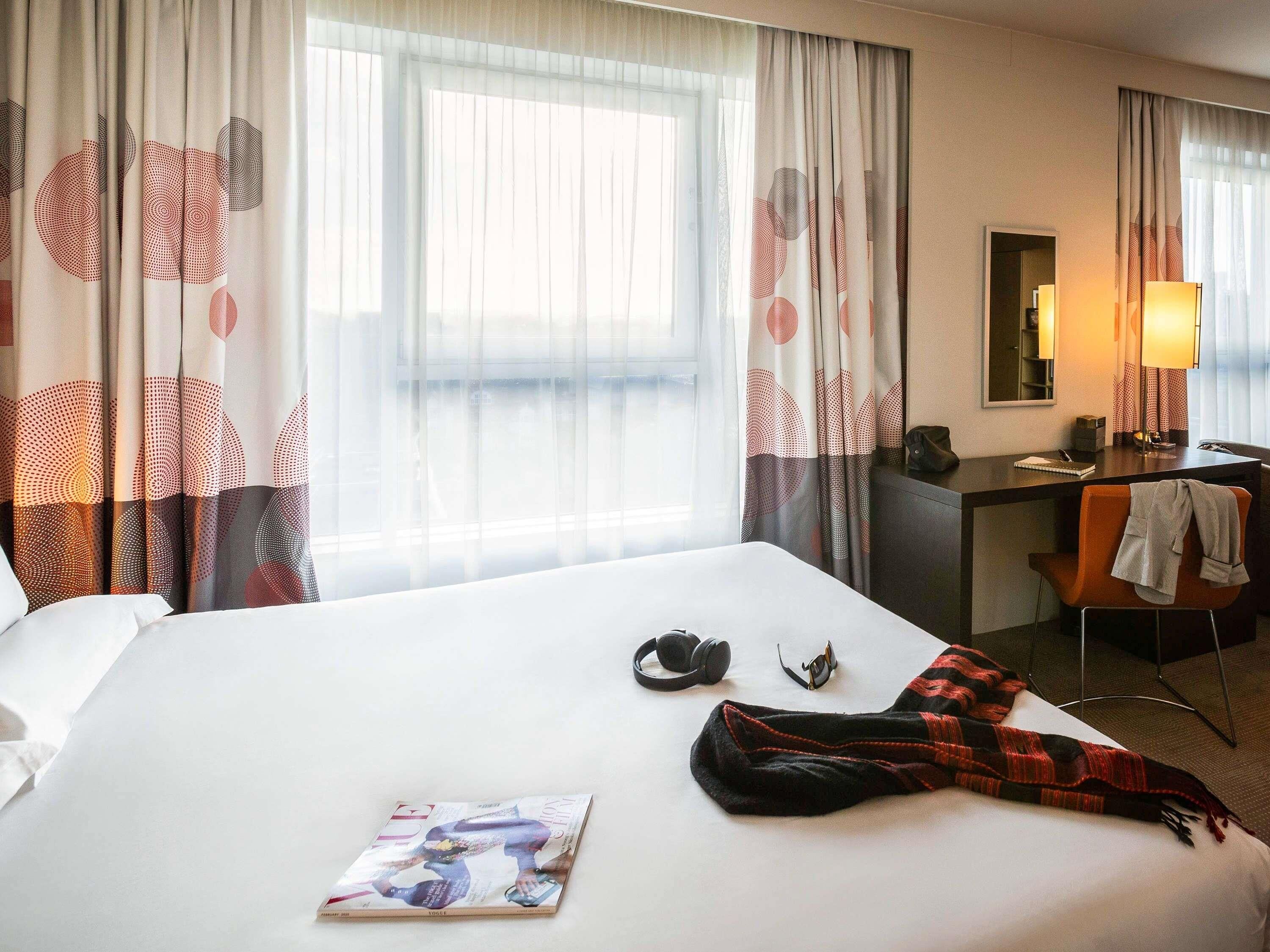 A hotel room with a bed, a desk, a chair, a lamp, and curtains with a circular pattern.