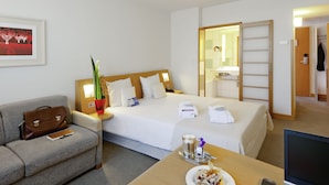 Habitación de hotel con cama, escritorio con ordenador portátil, mesa de comedor con comida, sofá, mesita de noche con lámpara y baño visible a través de una puerta abierta.