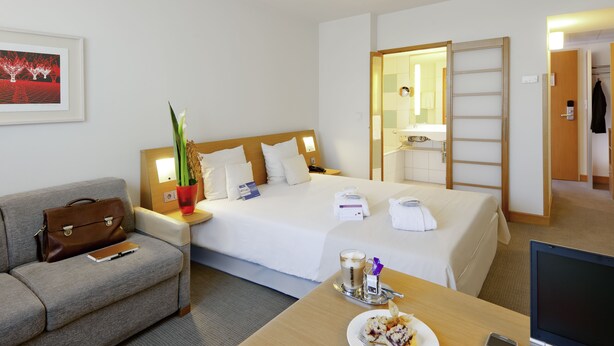 Habitación de hotel con cama, escritorio con ordenador portátil, mesa de comedor con comida, sofá, mesita de noche con lámpara y baño visible a través de una puerta abierta.