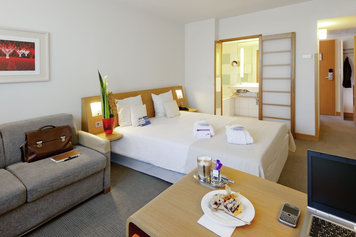 A hotel room with a bed, a desk with a laptop, a dining table with food, a sofa, a bedside table with a lamp, and a bathroom visible through an open door.