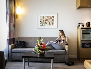 Una mujer sentada en un sofá gris en una habitación moderna con una máquina de café, una obra de arte abstracta enmarcada y un jarrón con flores sobre una mesa.