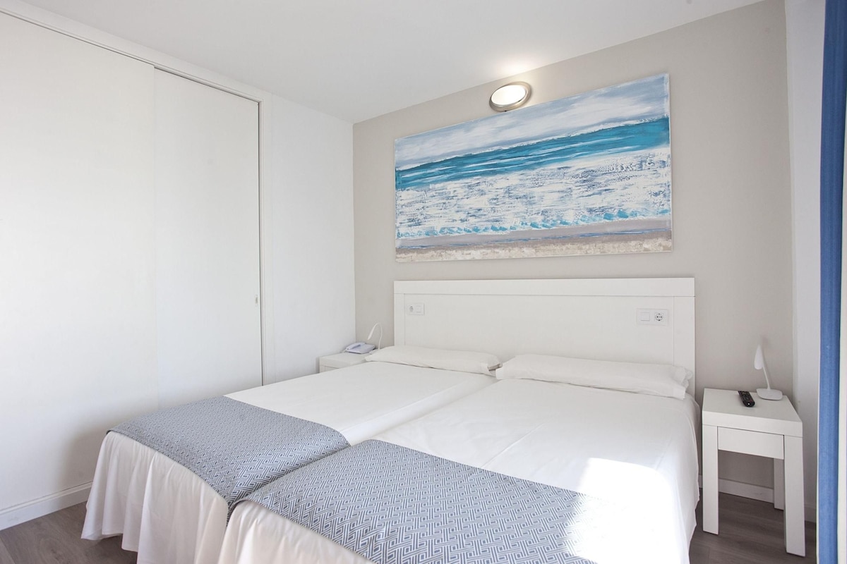 A double bed with a white headboard, a bedside table with a lamp, and a painting of a beach scene on the wall.