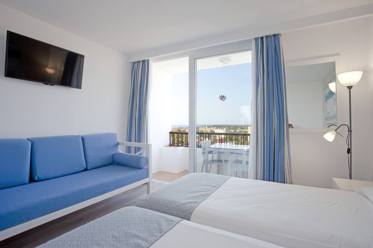 Habitación de hotel con cama, sofá azul, televisor, balcón con mesa y sillas, y una lámpara.