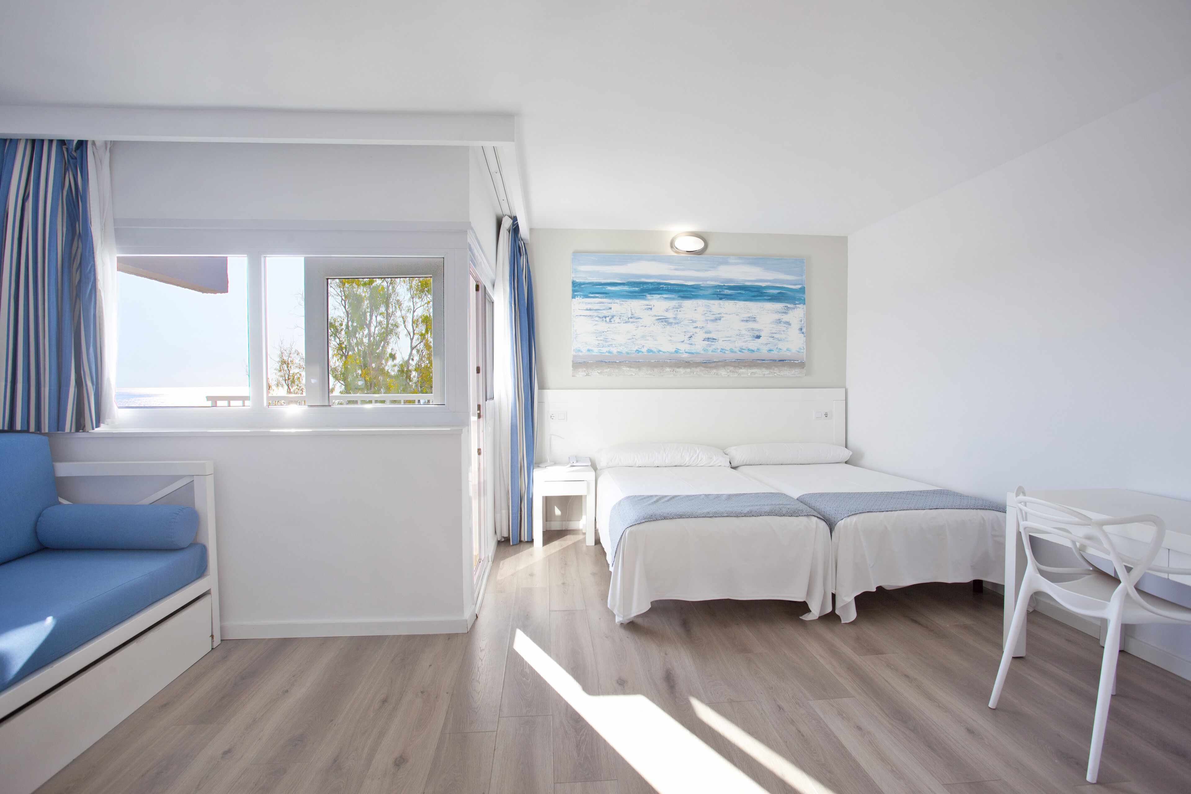 Un dormitorio con cama, una mesita, una silla, un ventanal con cortinas y un cuadro en la pared.