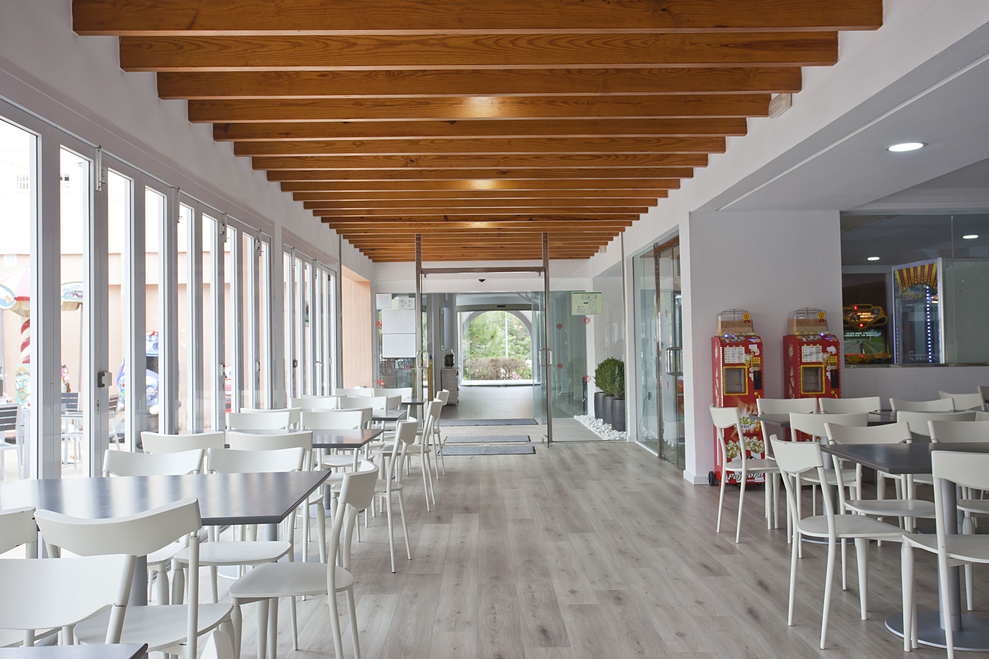 A modern interior with wooden ceiling beams, white tables and chairs, and large windows offering views of the outside.