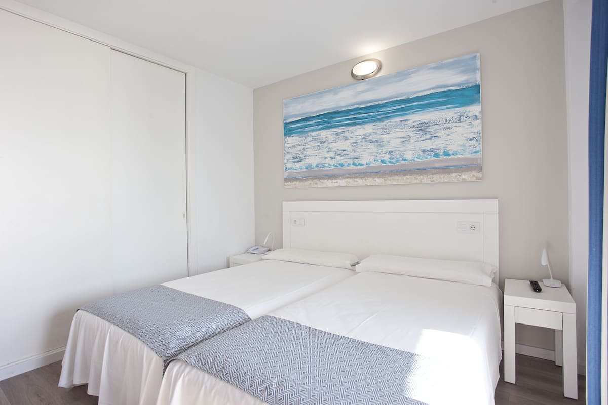 Una cama doble con cabecera blanca, una mesita de noche con lámpara y un cuadro colgado en la pared que representa una playa.
