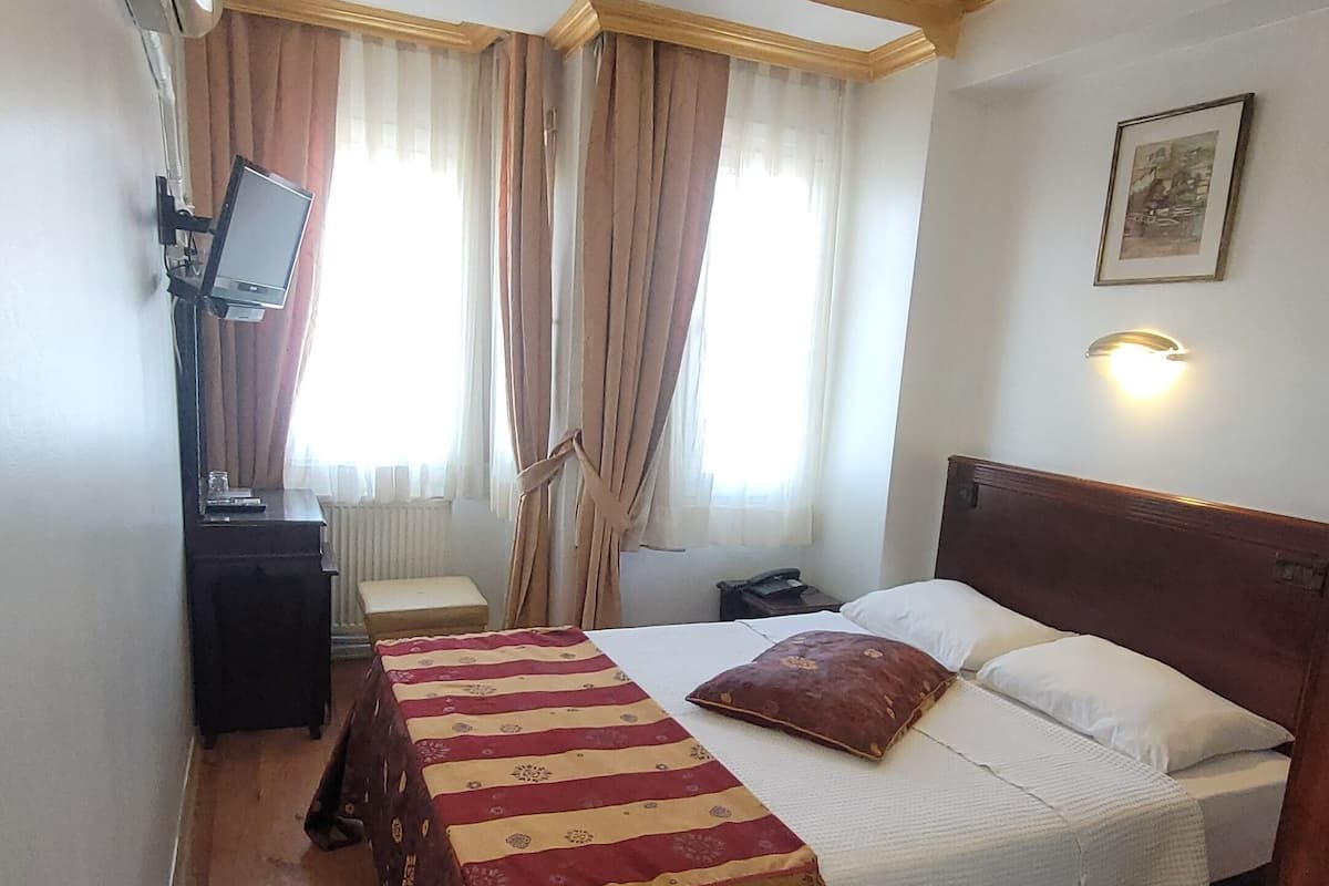 A hotel room with a bed, a desk, a chair, a radiator, a window with curtains, and a wall-mounted light fixture.