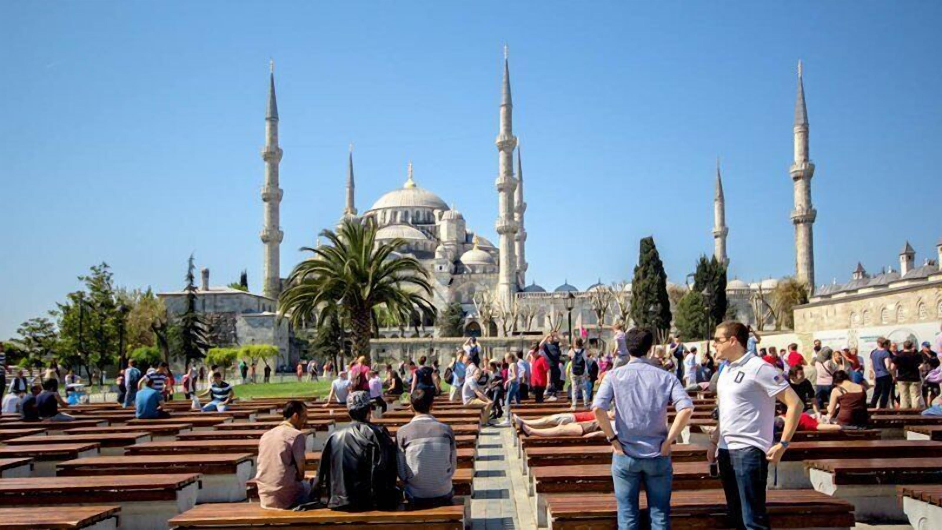 A mosque with multiple minarets and domes, surrounded by a courtyard with wooden benches and palm trees.