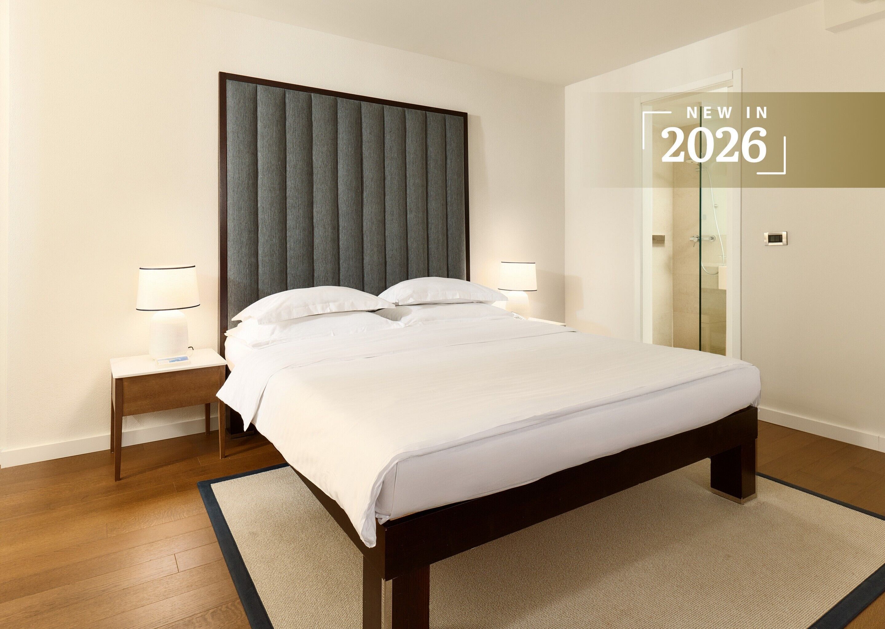 Una habitación de hotel moderna con una cama grande, dos mesitas de noche con lámparas, un box de ducha de vidrio y una paleta de colores neutros.