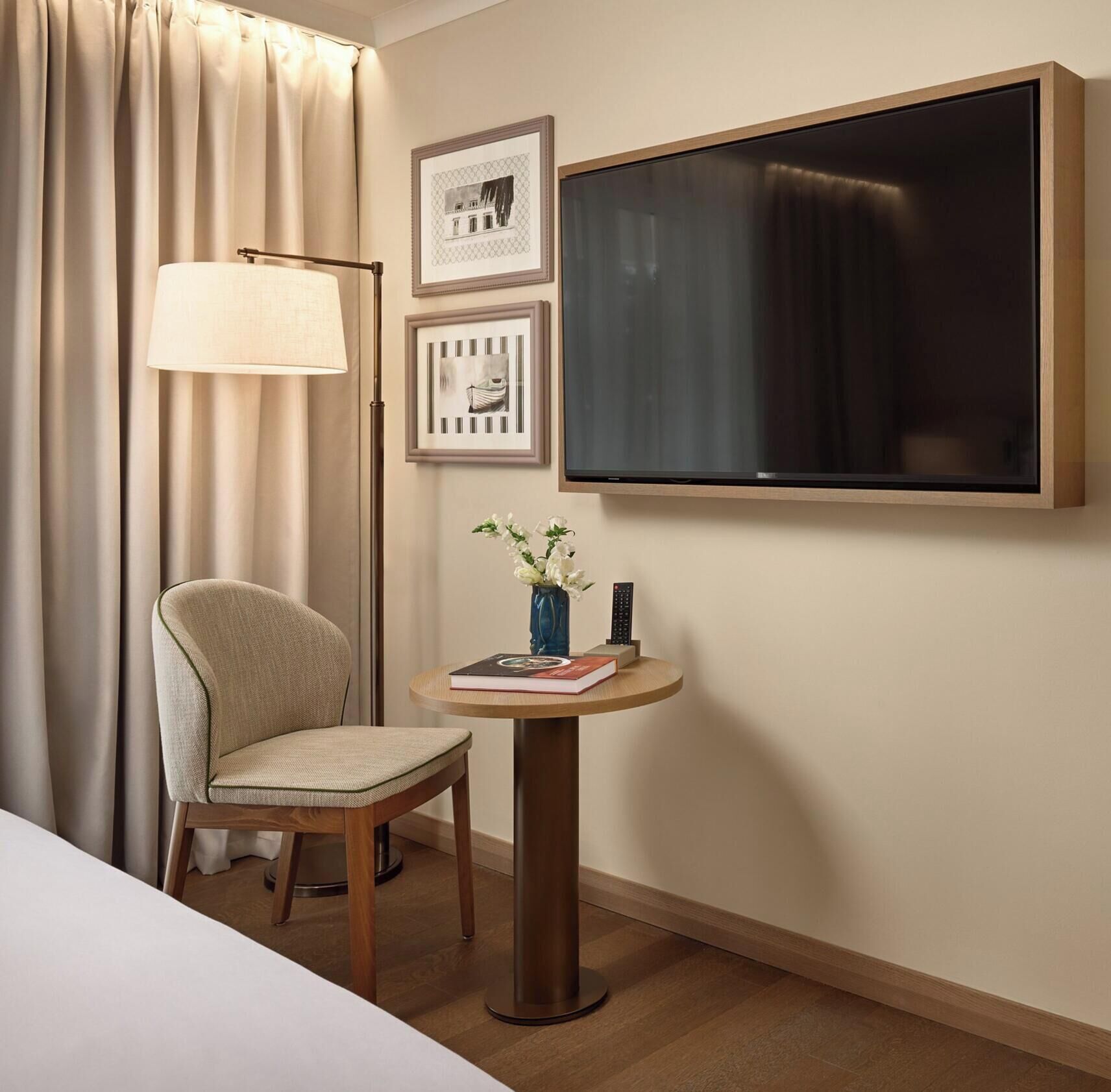 A hotel room with a bed, a chair, a small table with a vase and remote control, a lamp, and framed pictures on the wall.