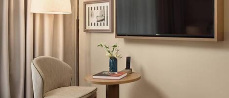 A hotel room with a bed, a chair, a small table with a vase and remote control, a lamp, and framed pictures on the wall.