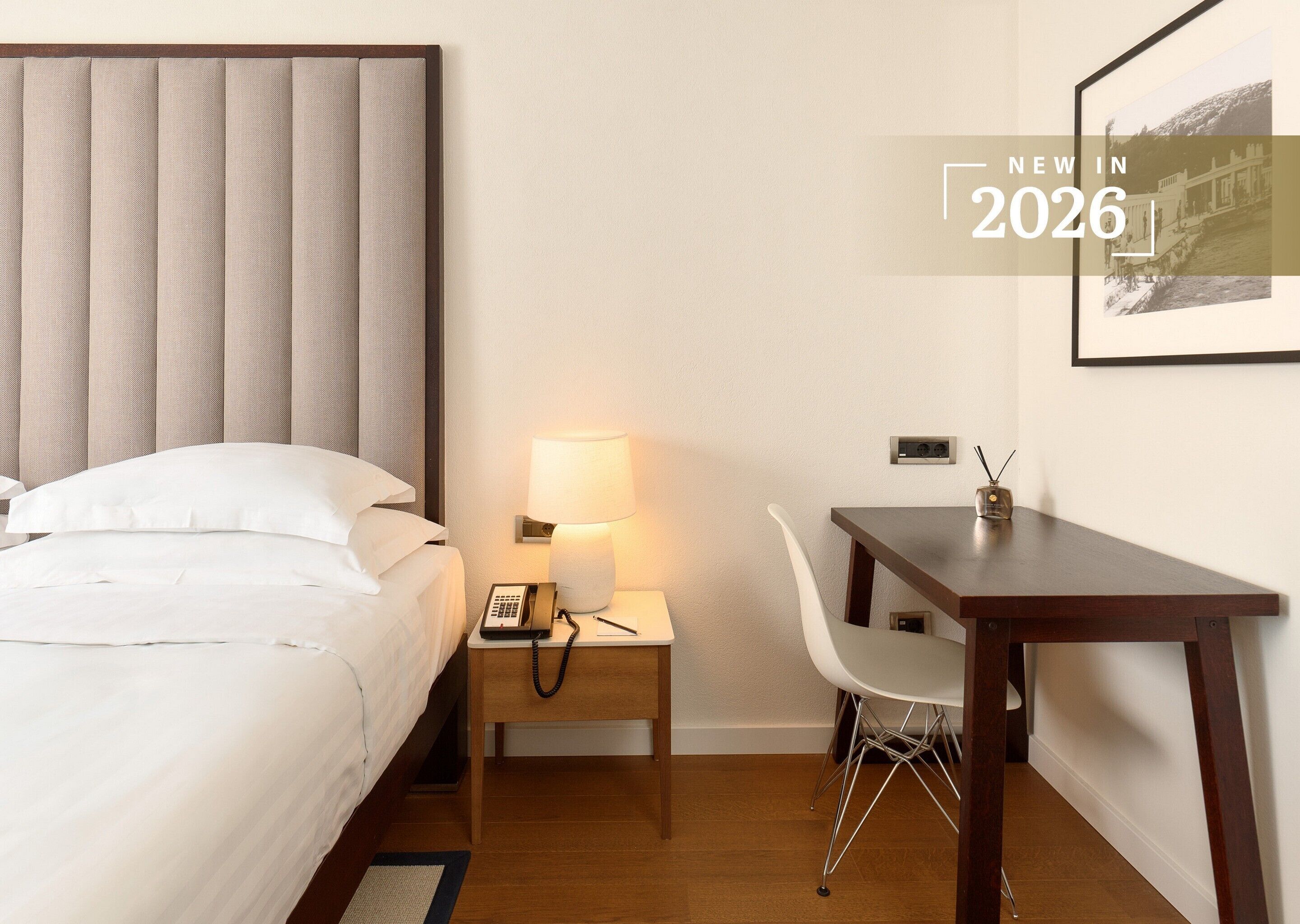 A hotel room with a bed, bedside table with a lamp, a desk with a chair, and a framed picture on the wall.